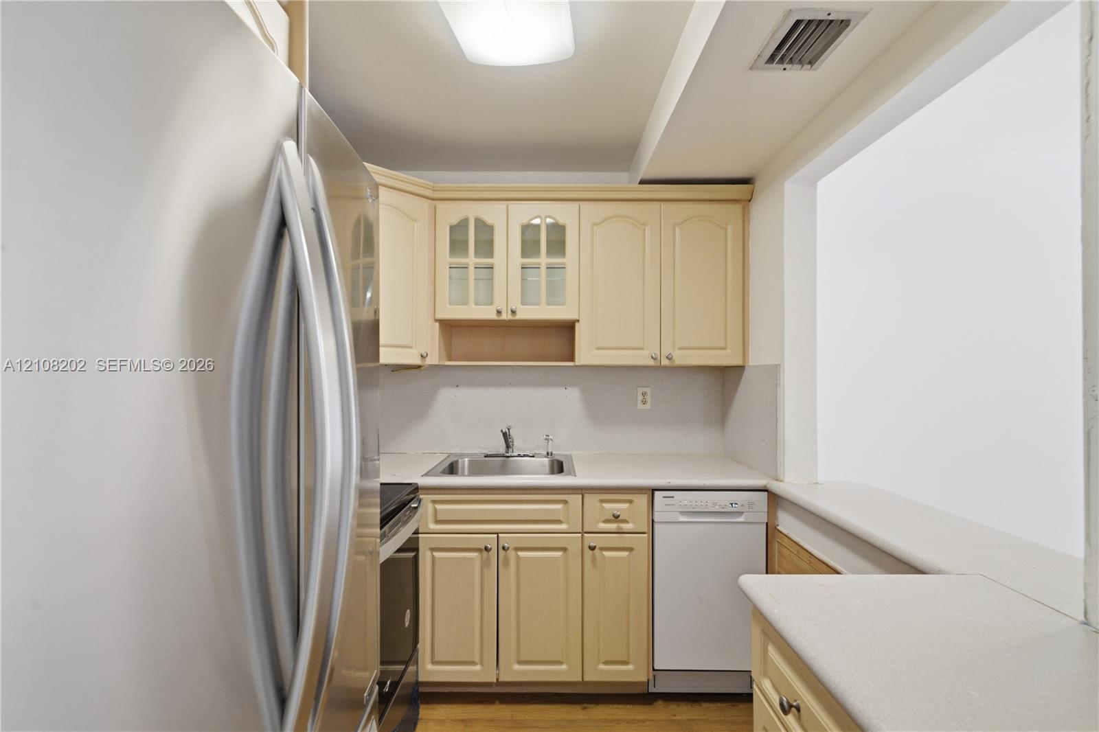Kitchen featuring light-tone cabinetry with glass-paneled upper doors, solid-surface countertops, an integrated stainless steel sink, and a built-in dishwasher Kitchen featuring light-tone cabinetry with glass-paneled upper doors, solid-surface countertops, an integrated stainless steel sink, and a built-in dishwasher