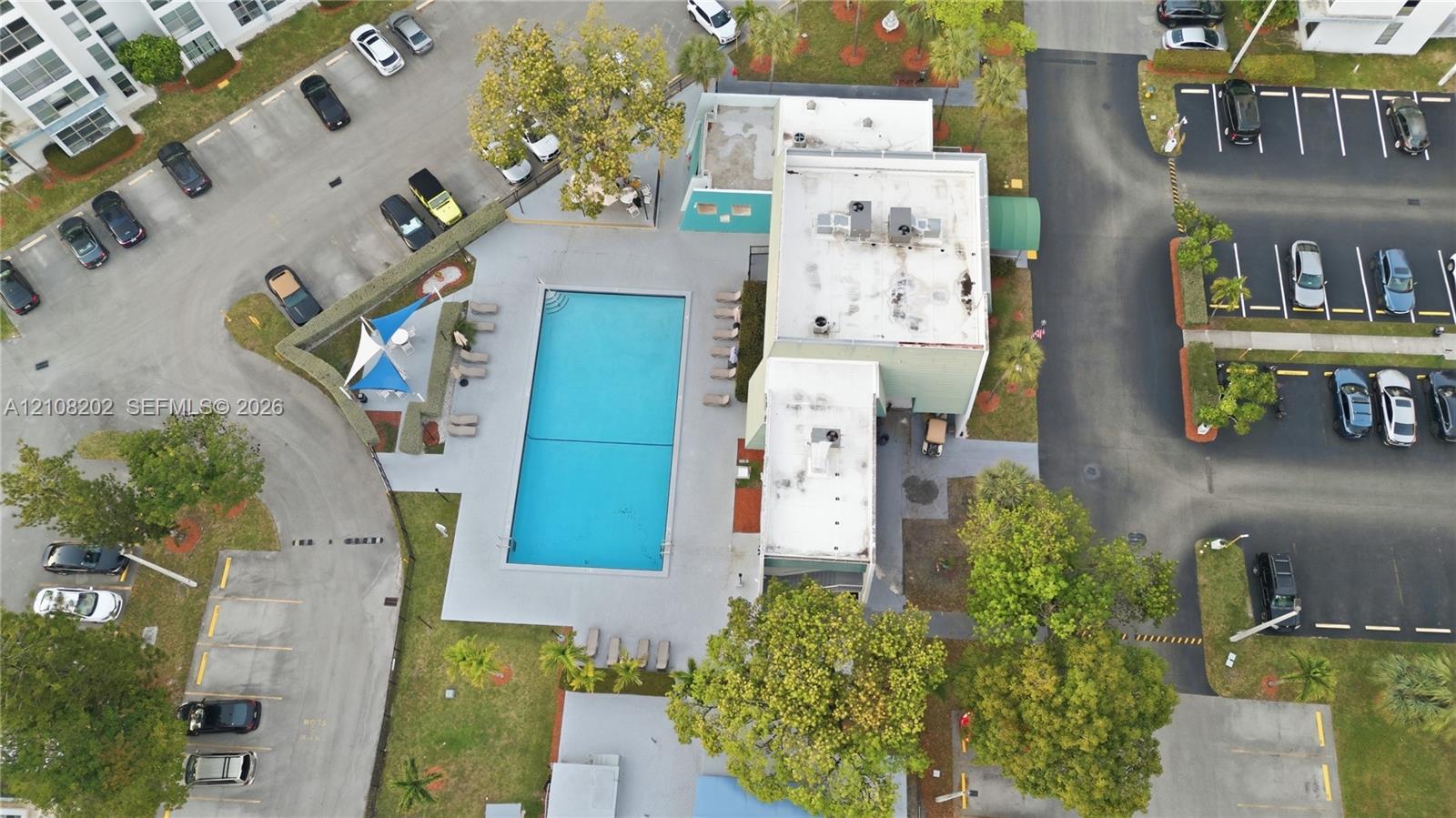 Aerial perspective of the community recreation area featuring a swimming pool, paved pool deck, shade structures, and surrounding landscaped grounds with established trees