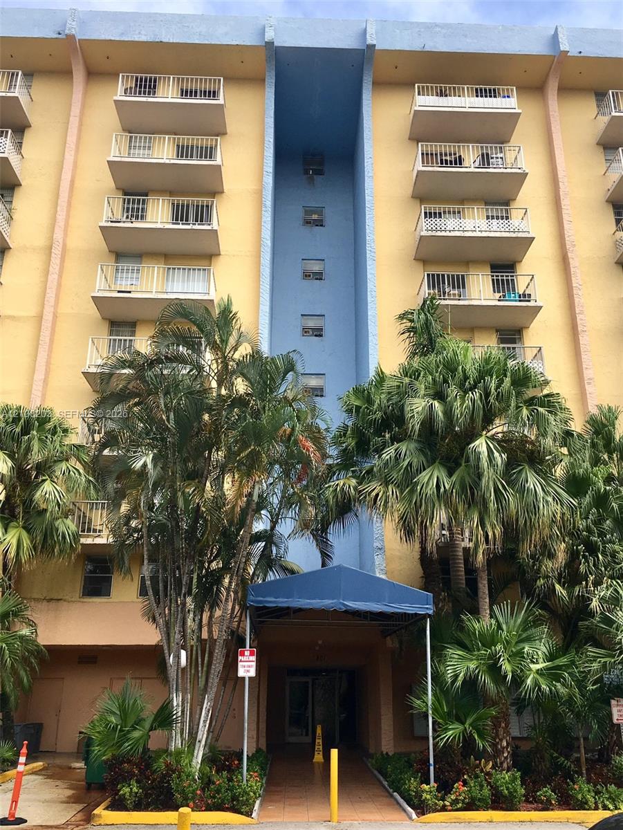 Multi-story residential building exterior featuring private balconies with white railings, a covered entryway with a blue awning, and tropical landscaping