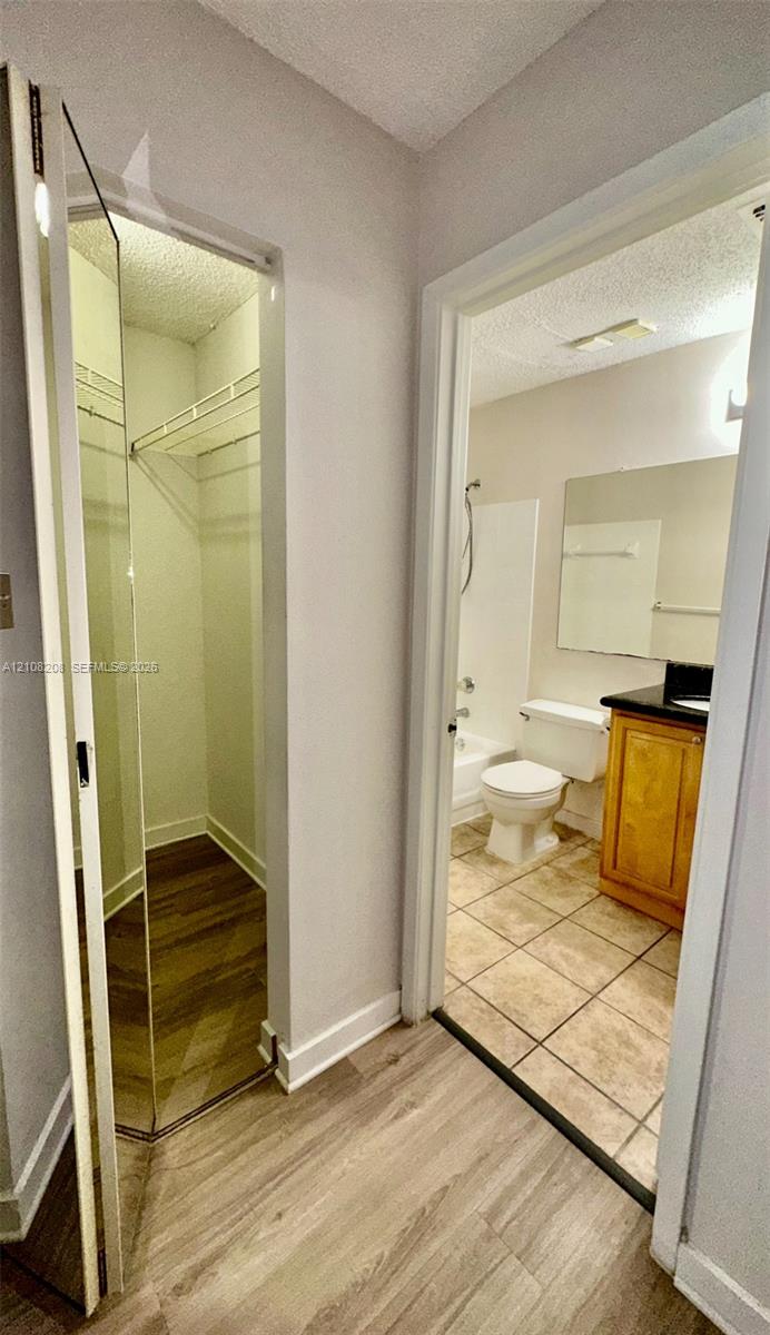 Walk-in closet with wire shelving and wood-finish flooring, featuring a bathroom with a wood-finish vanity cabinet, stone countertop, ceramic tile flooring, and a shower-tub combination