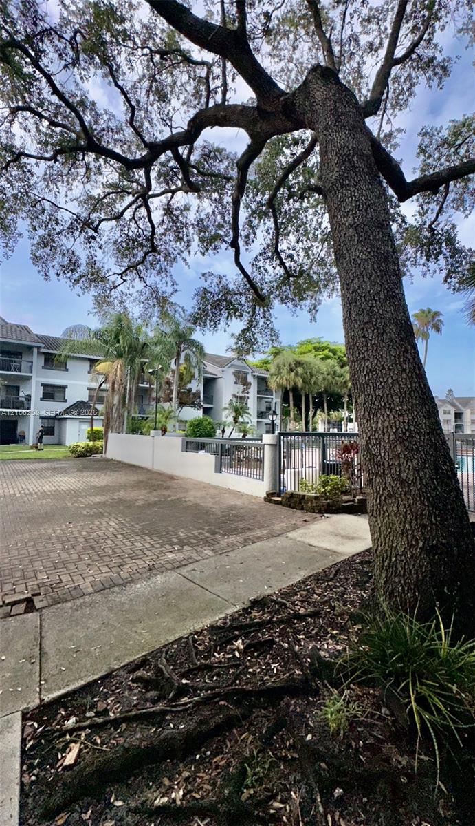 Exterior grounds featuring paved brick surfacing, mature canopy trees, concrete walkways, and metal perimeter fencing