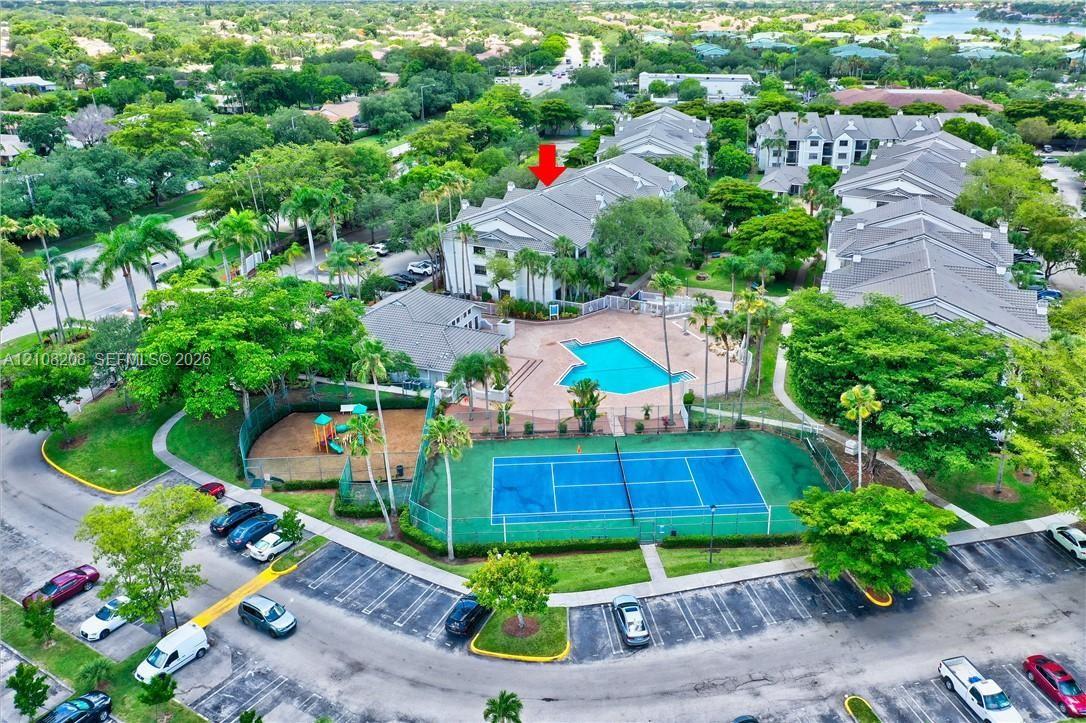 Multi-story building featuring a pitched roof, community swimming pool, fenced tennis court, and lush mature landscaping
