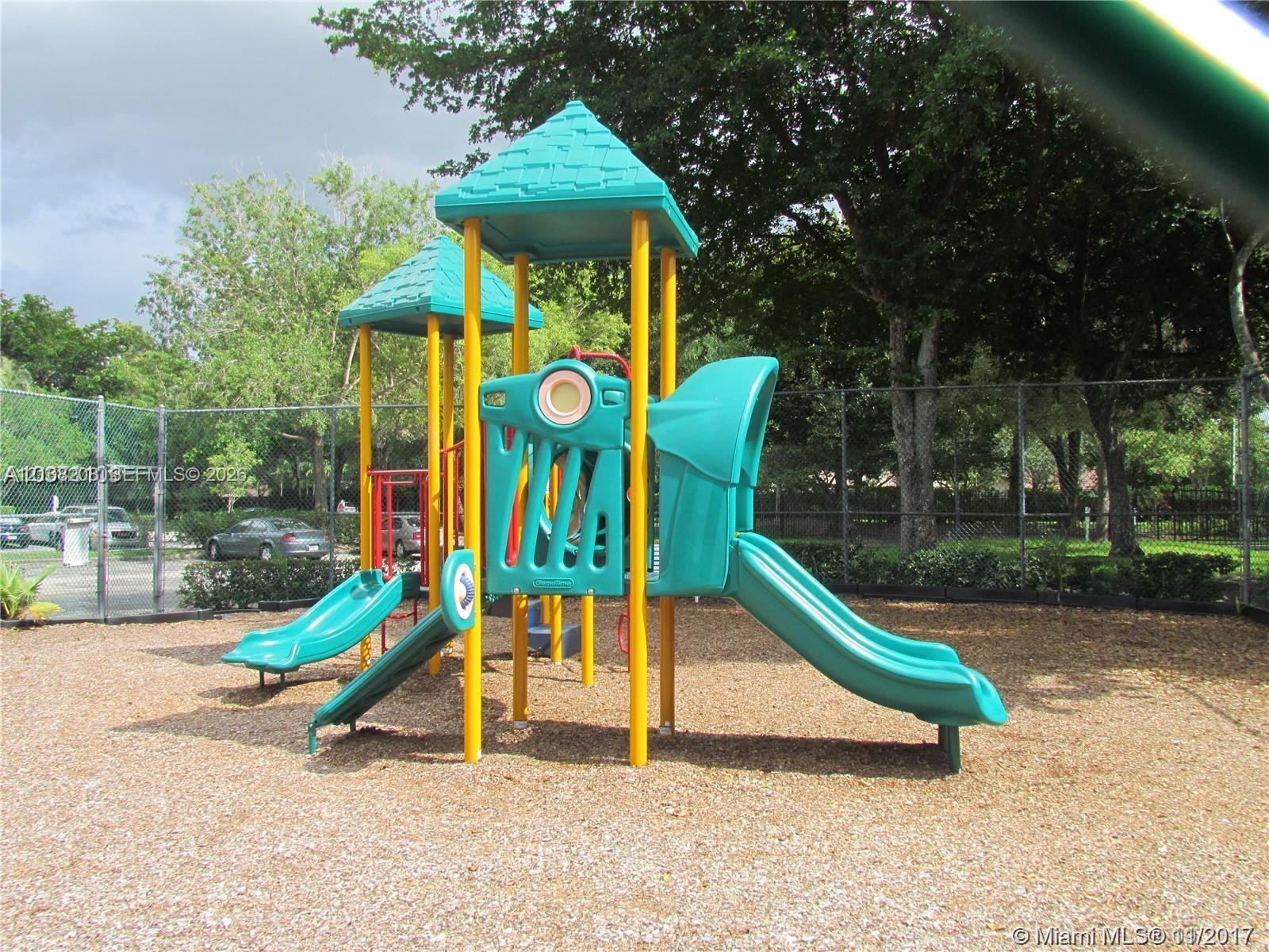 Outdoor recreational structure featuring a multi-level composite play system, dual slides, and wood-chip ground covering, enclosed by chain-link fencing