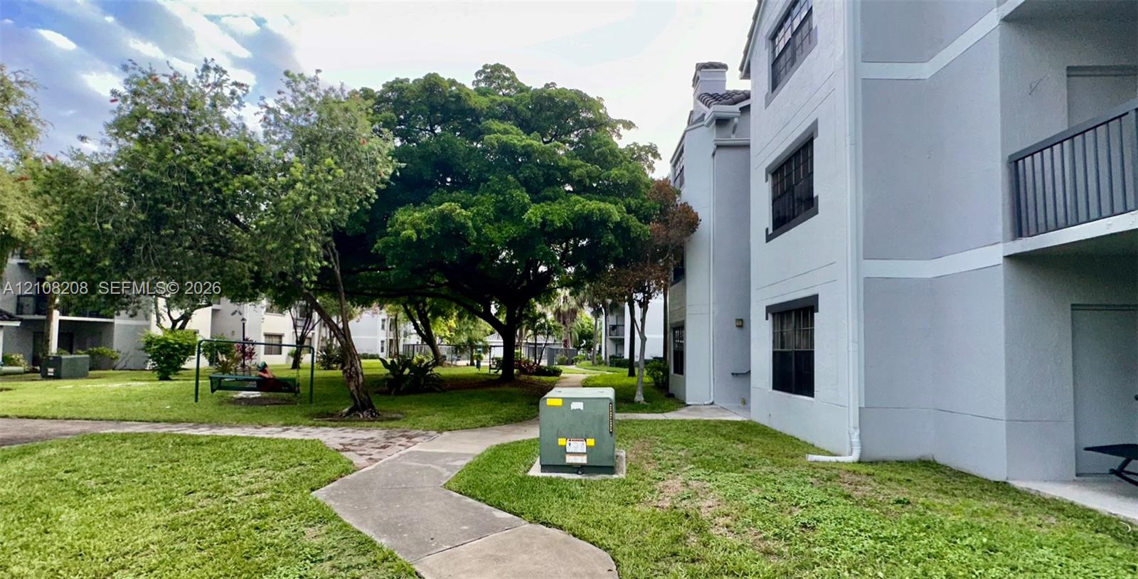 Multi-story exterior featuring a light gray facade, dark-framed windows, and private balconies, set among mature trees and manicured lawn areas with concrete pathways