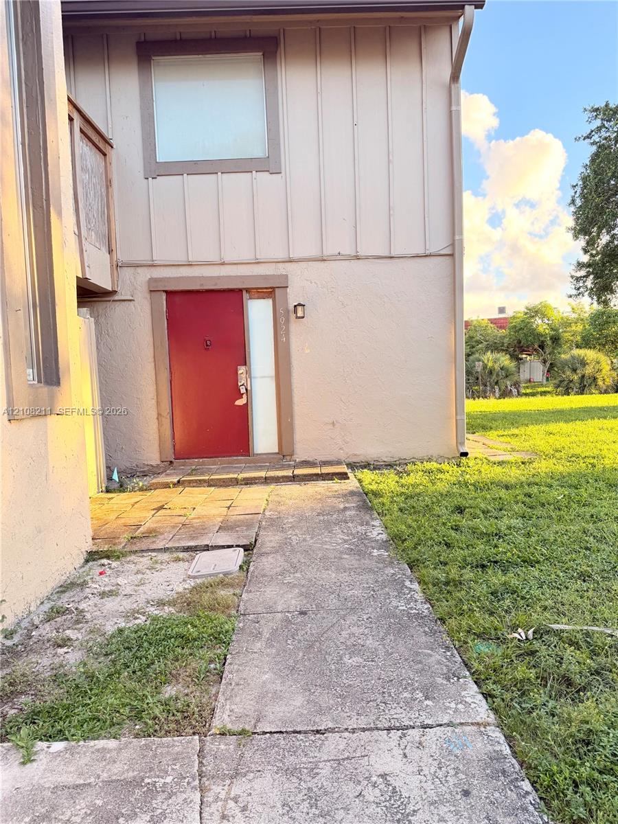 Exterior featuring vertical board-and-batten siding, stucco finish, a red entry door with sidelight, and a concrete paver walkway