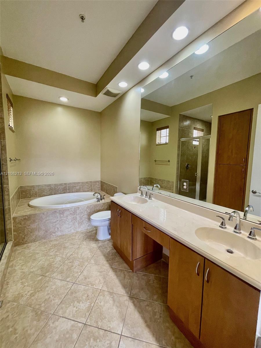 Dual-sink vanity with stone-finish countertop, soaking tub surrounded by tile, full-width mirror, recessed lighting, and tile flooring