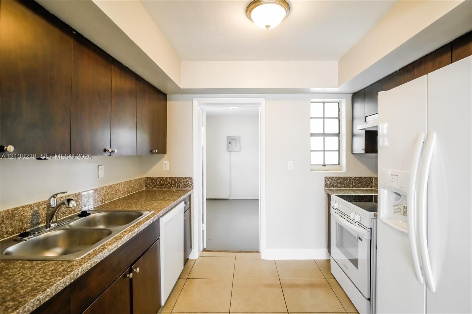 Kitchen featuring dark wood-finish cabinetry, stone-surface countertops, double-basin stainless steel sink, ceramic tile flooring, and integrated white appliances Kitchen featuring dark wood-finish cabinetry, stone-surface countertops, double-basin stainless steel sink, ceramic tile flooring, and integrated white appliances