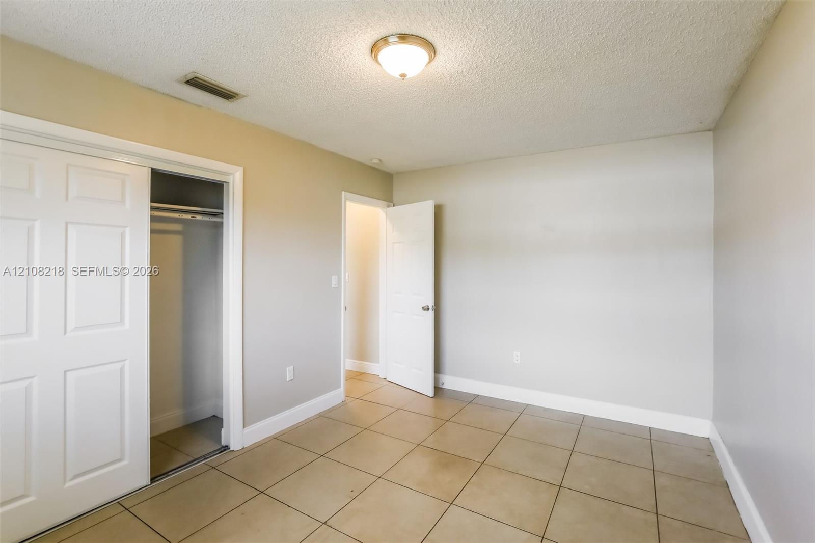 Neutral-toned room featuring large-format square tile flooring, a sliding bypass closet door, a solid-panel door, and a flush-mount ceiling light fixture Neutral-toned room featuring large-format square tile flooring, a sliding bypass closet door, a solid-panel door, and a flush-mount ceiling light fixture
