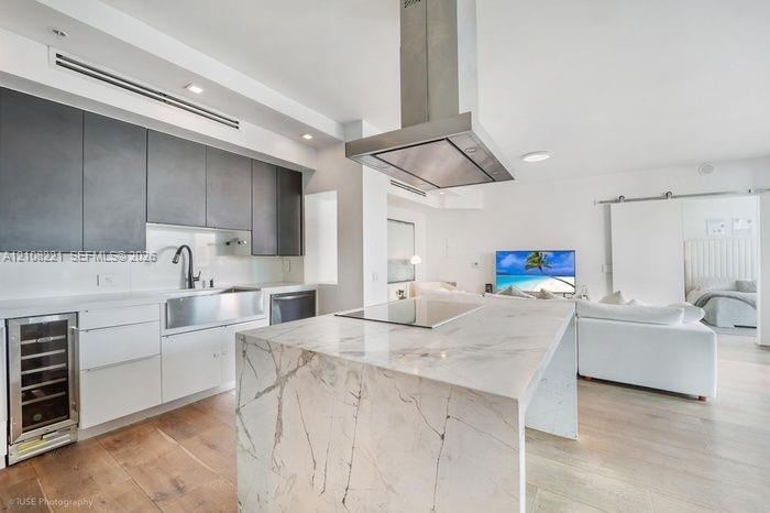 Kitchen featuring a waterfall-edge island with stone surfacing, stainless steel farmhouse sink, matte finish upper cabinetry, integrated wine refrigerator, and stainless steel overhead range hood