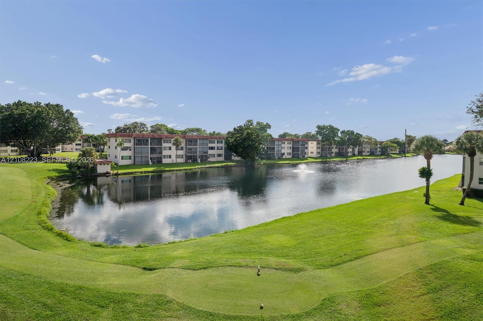 Waterfront landscape featuring a pond, fountain, manicured lawn, mature trees, and palm trees