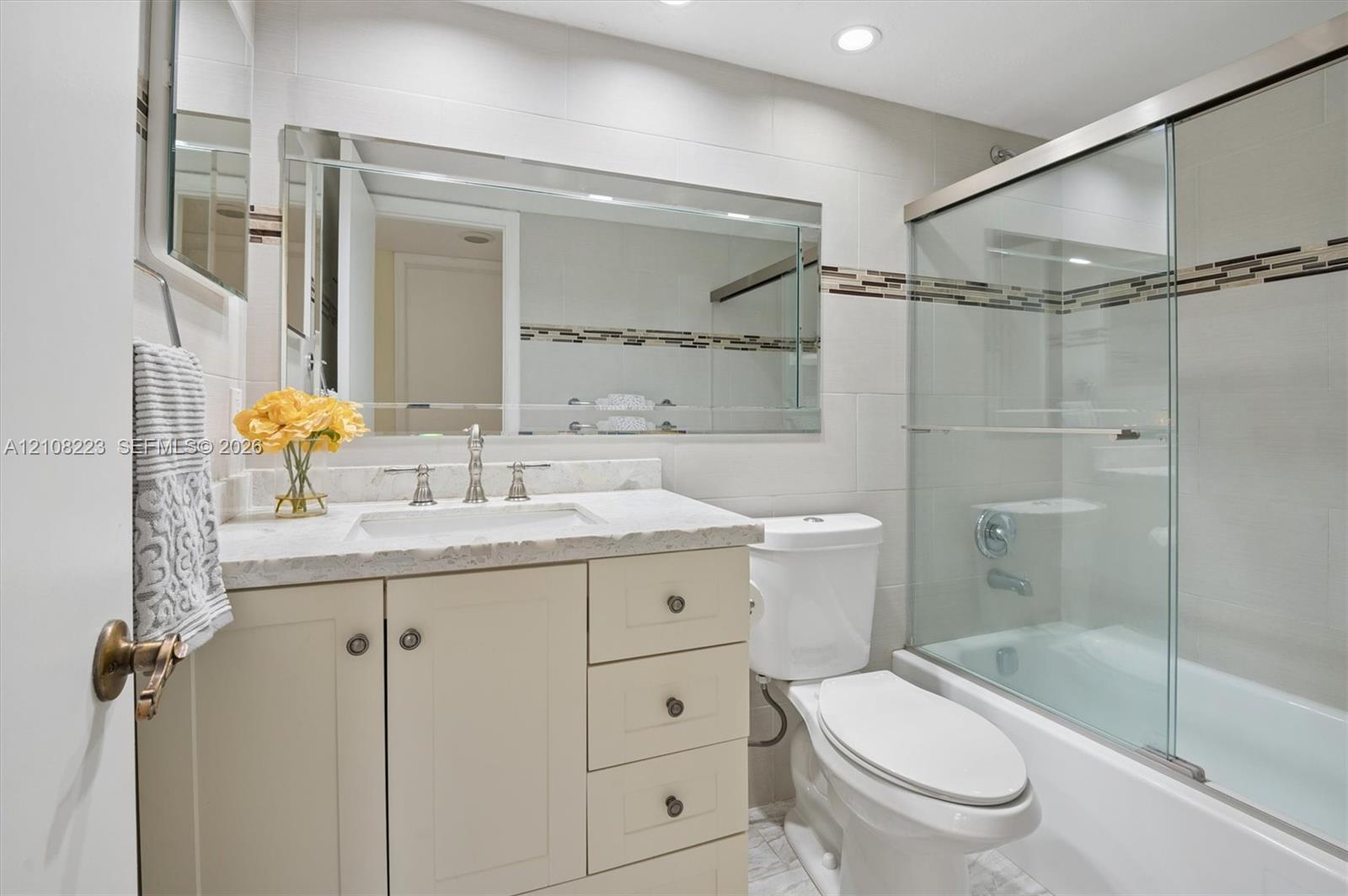 Bathroom featuring a vanity with stone-finish countertop, undermount sink, recessed lighting, and a bathtub with glass sliding doors and decorative tile inlay