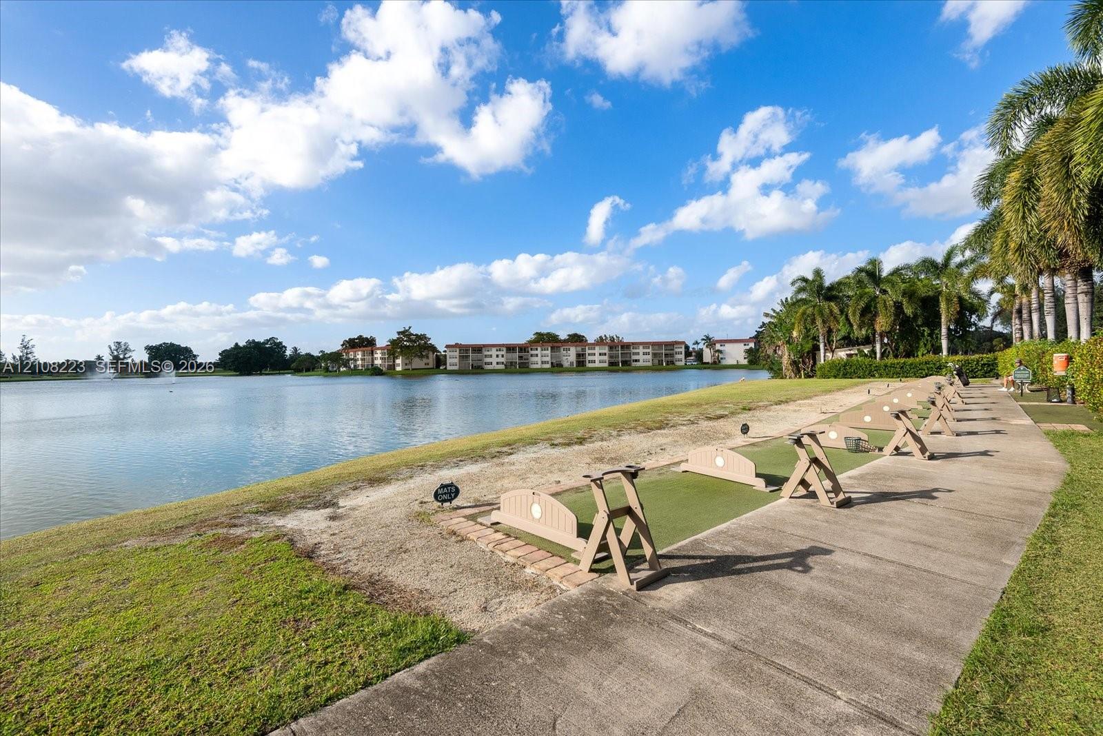 Waterfront driving range featuring a concrete walkway, designated tee boxes with synthetic turf, and mature palm trees