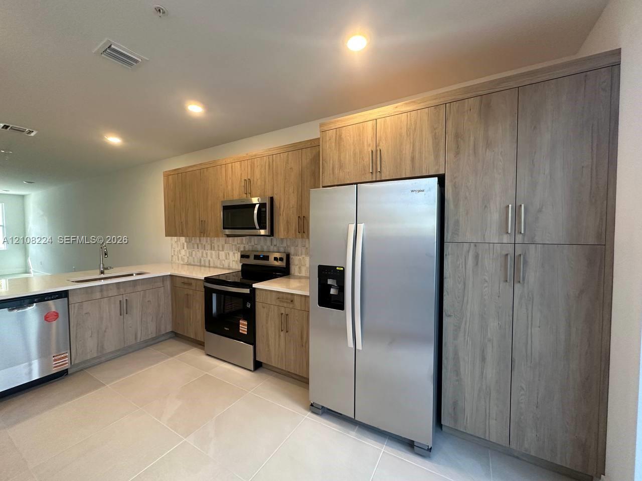 Kitchen featuring textured wood-finish cabinetry, stainless steel appliances, light-toned solid surface countertops, a decorative mosaic tile backsplash, and large-format floor tile