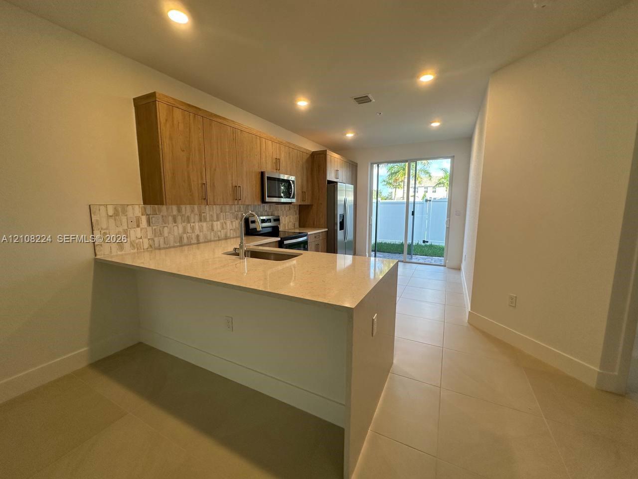 Kitchen featuring wood-finish cabinetry, quartz countertops, a stone-tile backsplash, stainless steel appliances, and large-format tile flooring Kitchen featuring wood-finish cabinetry, quartz countertops, a stone-tile backsplash, stainless steel appliances, and large-format tile flooring
