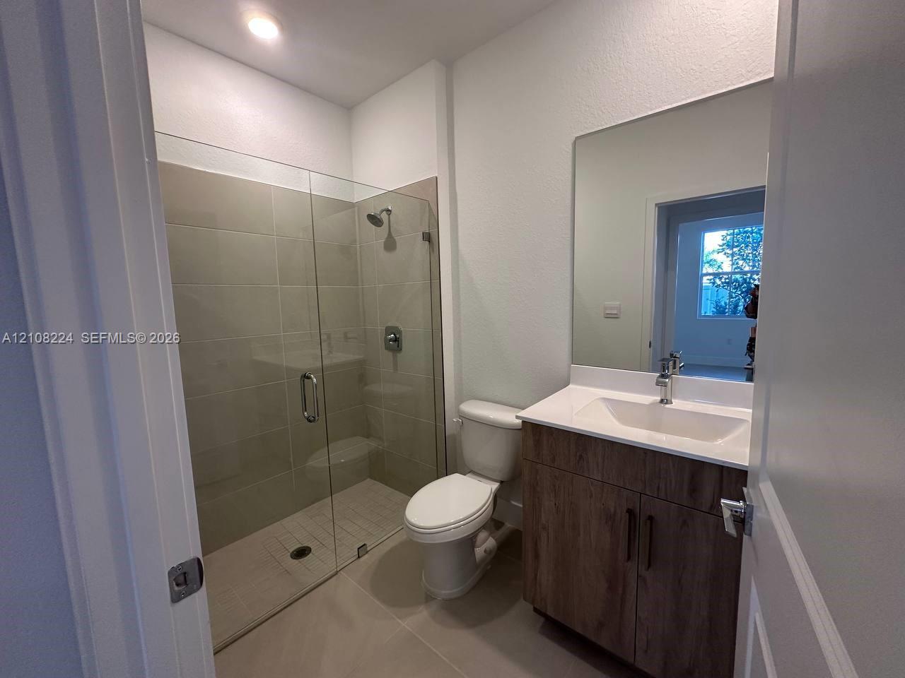 Walk-in shower with floor-to-ceiling tile, wood-finish vanity with an integrated basin, countertop with stone-style surface, and frameless vanity mirror Walk-in shower with floor-to-ceiling tile, wood-finish vanity with an integrated basin, countertop with stone-style surface, and frameless vanity mirror