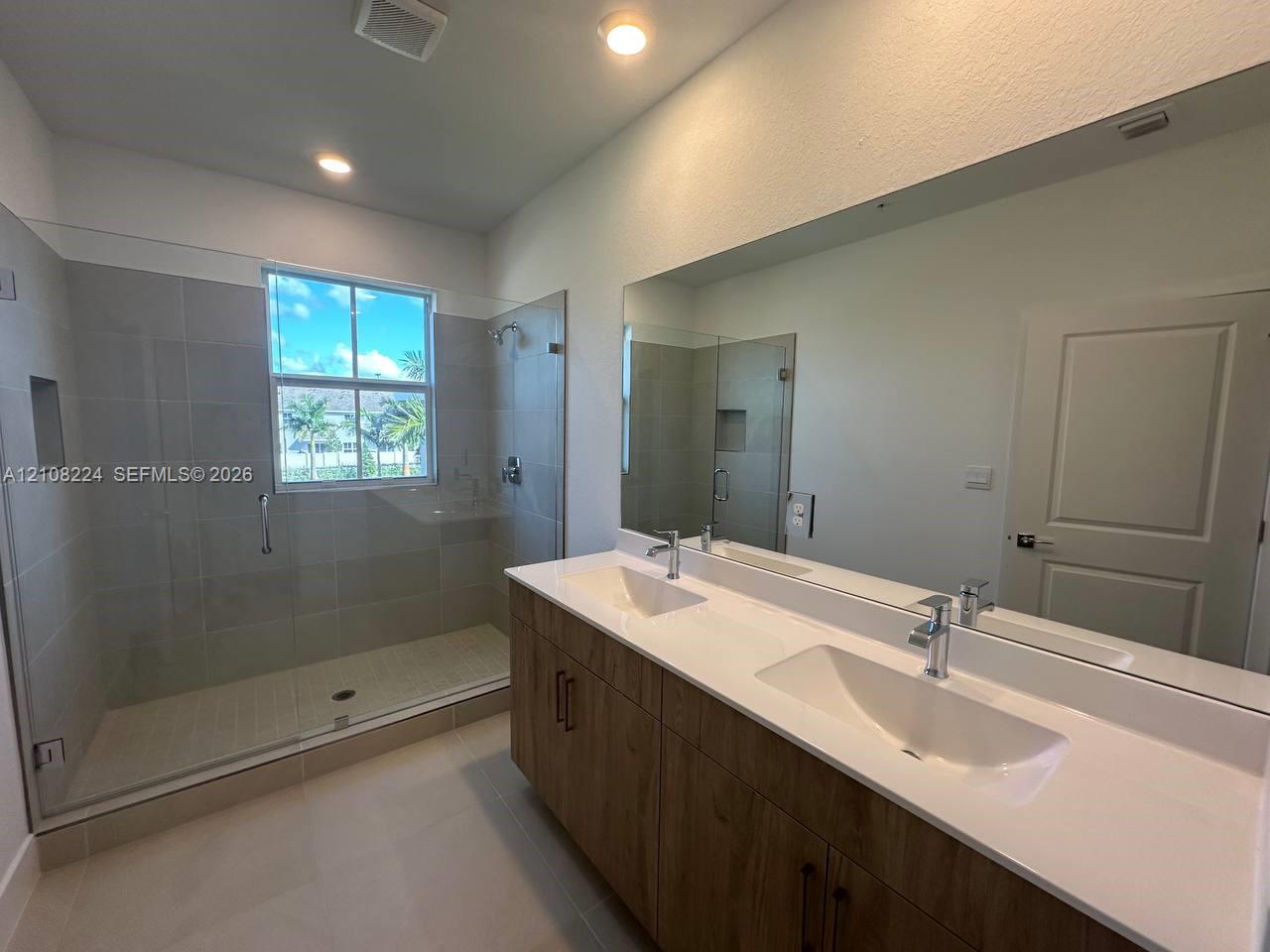 Dual-sink vanity with a white countertop and wood-finish cabinetry, frameless glass-enclosed shower featuring tile walls, integrated shower niche, and a large wall-mounted mirror