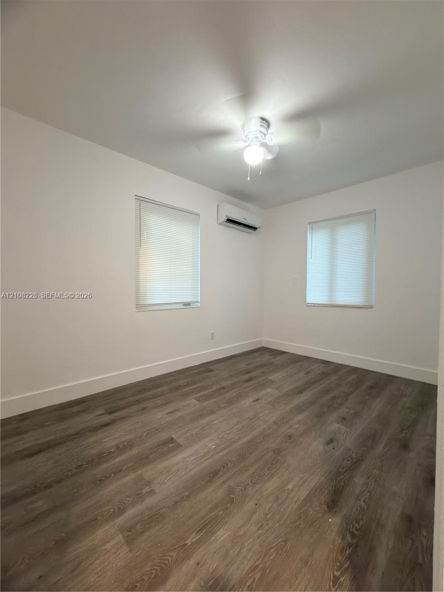 Interior space featuring wood-finish flooring, two dual-pane windows, a wall-mounted climate control unit, and a ceiling-mounted light fixture with fan