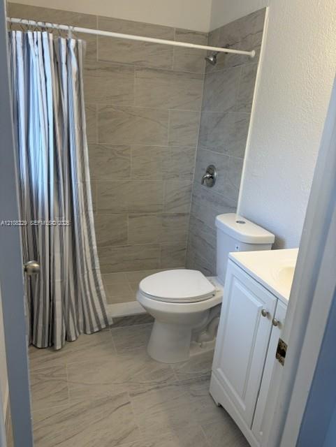 Bathroom featuring large-format tile flooring, a shower surround with neutral tile detailing, a white vanity cabinet, and a two-piece toilet