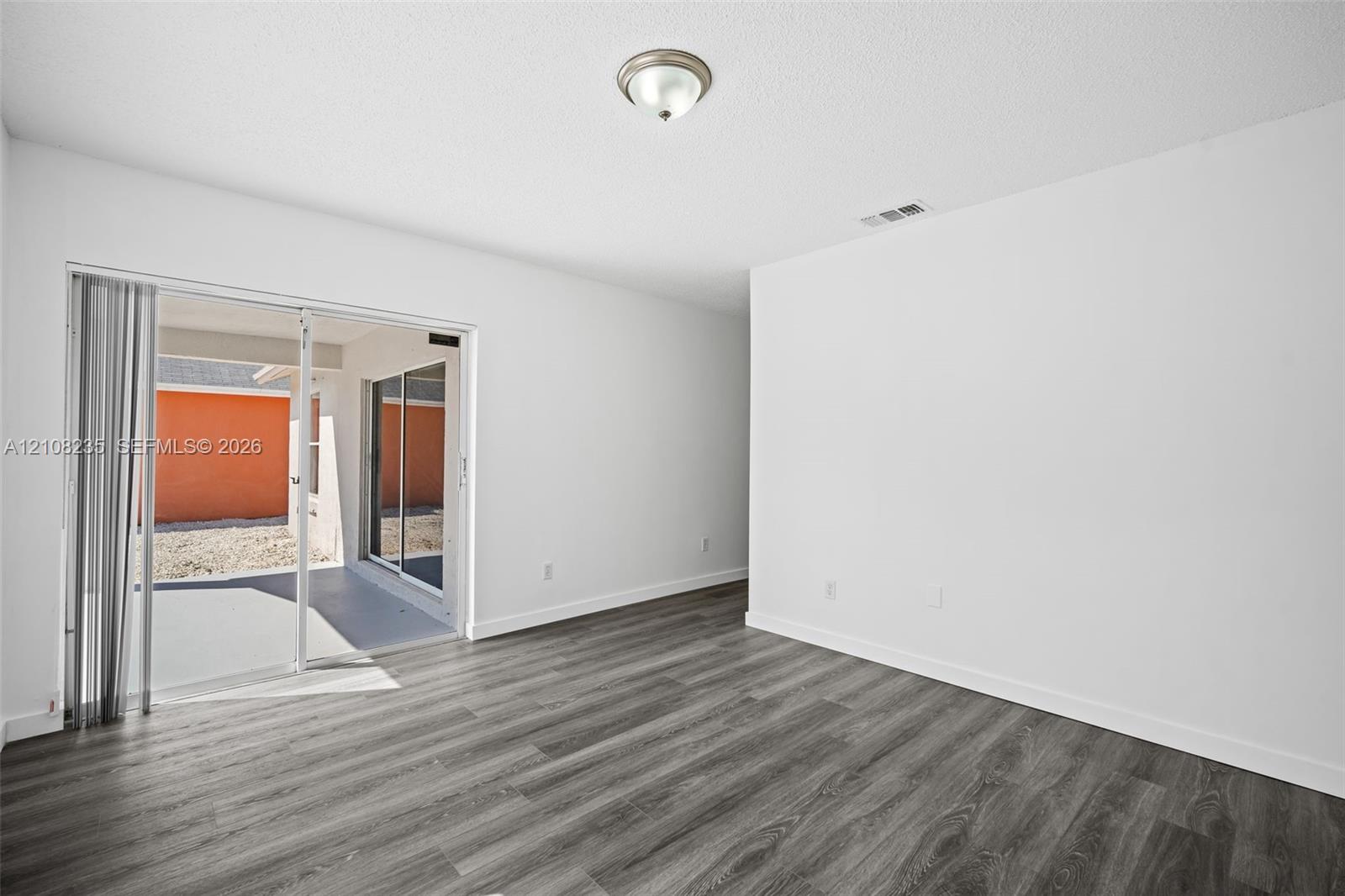 Interior space featuring wood-finish flooring, white walls, flush-mount ceiling lighting, and sliding glass doors with vertical blinds leading to a paved patio area