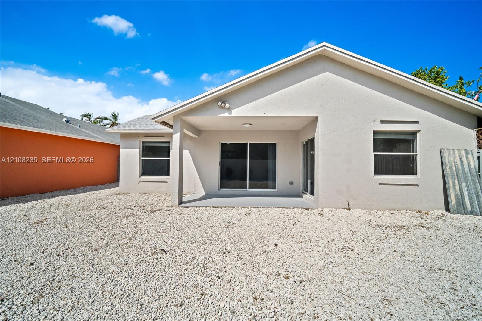 Rear exterior elevation featuring a stucco facade, covered patio, sliding glass doors, and gravel landscaping