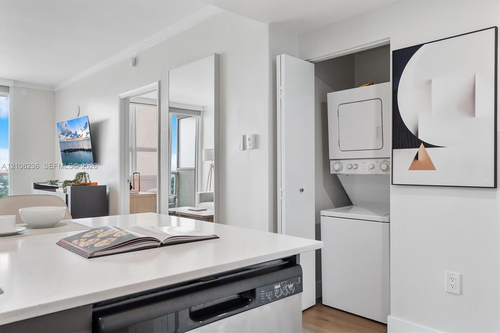 Kitchen featuring a white solid-surface countertop island, integrated stainless steel dishwasher, and stacked laundry unit housed in a bi-fold door closet