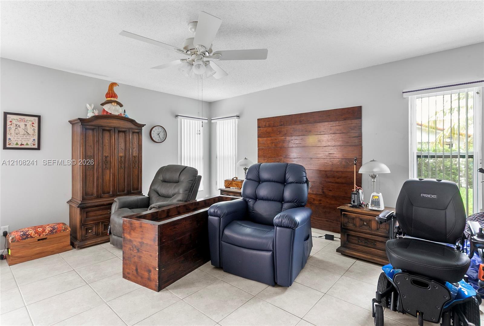 Room interior featuring light-toned tile flooring, wood-paneled accent wall, dual windows with vertical blinds, and a white ceiling fan with integrated lighting Room interior featuring light-toned tile flooring, wood-paneled accent wall, dual windows with vertical blinds, and a white ceiling fan with integrated lighting