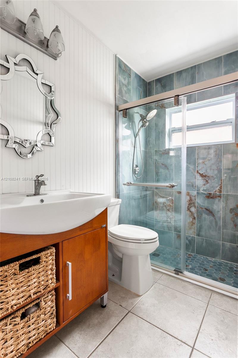 Vanity with integrated sink and open shelving, beadboard wall paneling, shower enclosure with sliding glass door, multi-tonal tiled shower surround, and hexagonal floor tile
