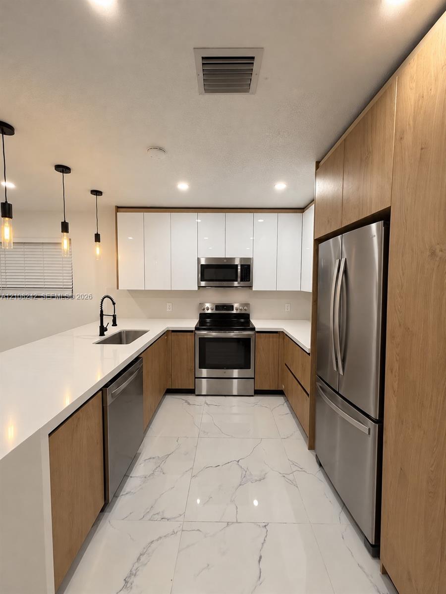 Kitchen featuring white gloss and wood-finish cabinetry, white solid-surface countertops, large-format veined tile flooring, stainless steel appliances, and matte black pendant lighting