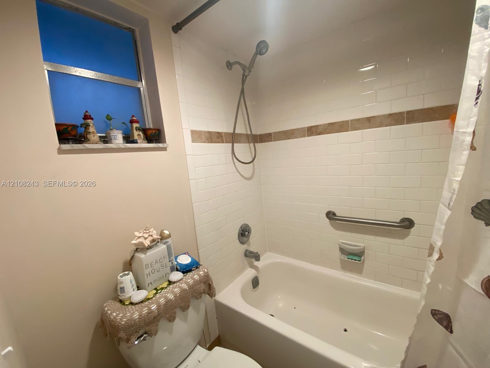 Bathtub with white subway tile surround, decorative tile inlay, adjustable shower head, and wall-mounted grab bar, complemented by a stone-finish window sill