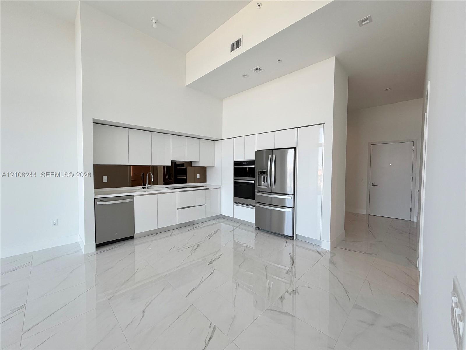 Kitchen featuring high-gloss white cabinetry, stainless steel appliances, built-in wall oven, metallic backsplash, and large-format porcelain tile flooring
