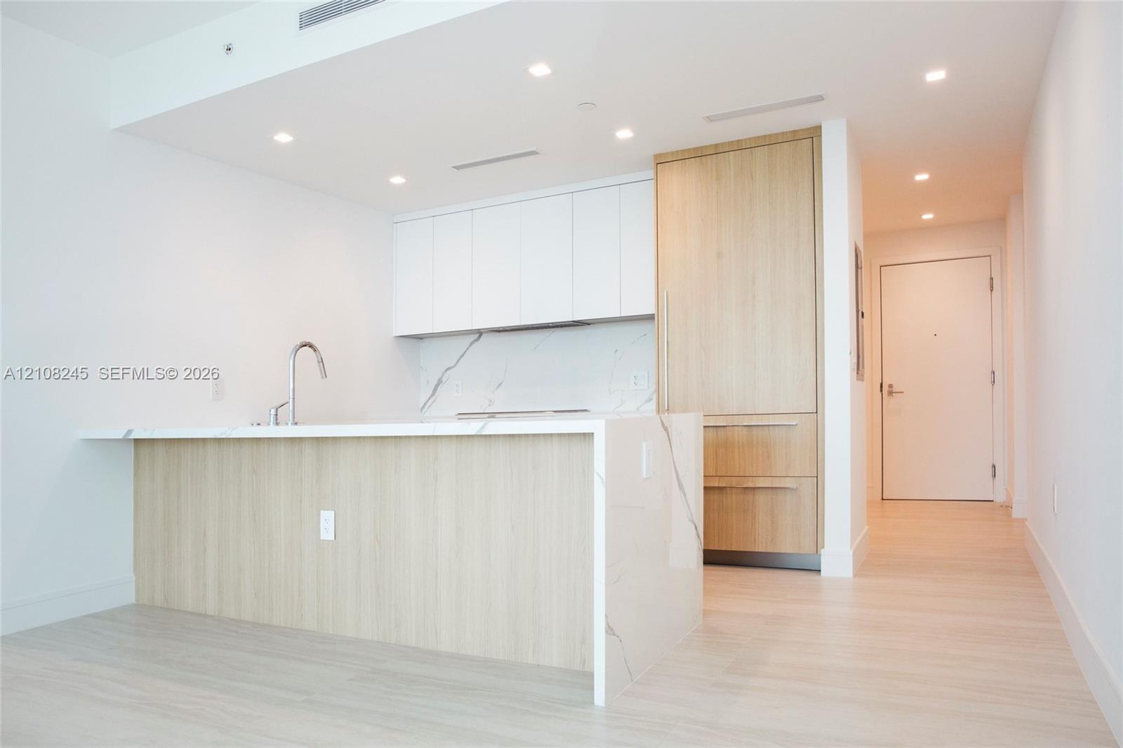 Kitchen featuring wood-finish cabinetry, white matte upper cabinets, stone-surfaced island with waterfall edge, integrated refrigerator, and recessed lighting