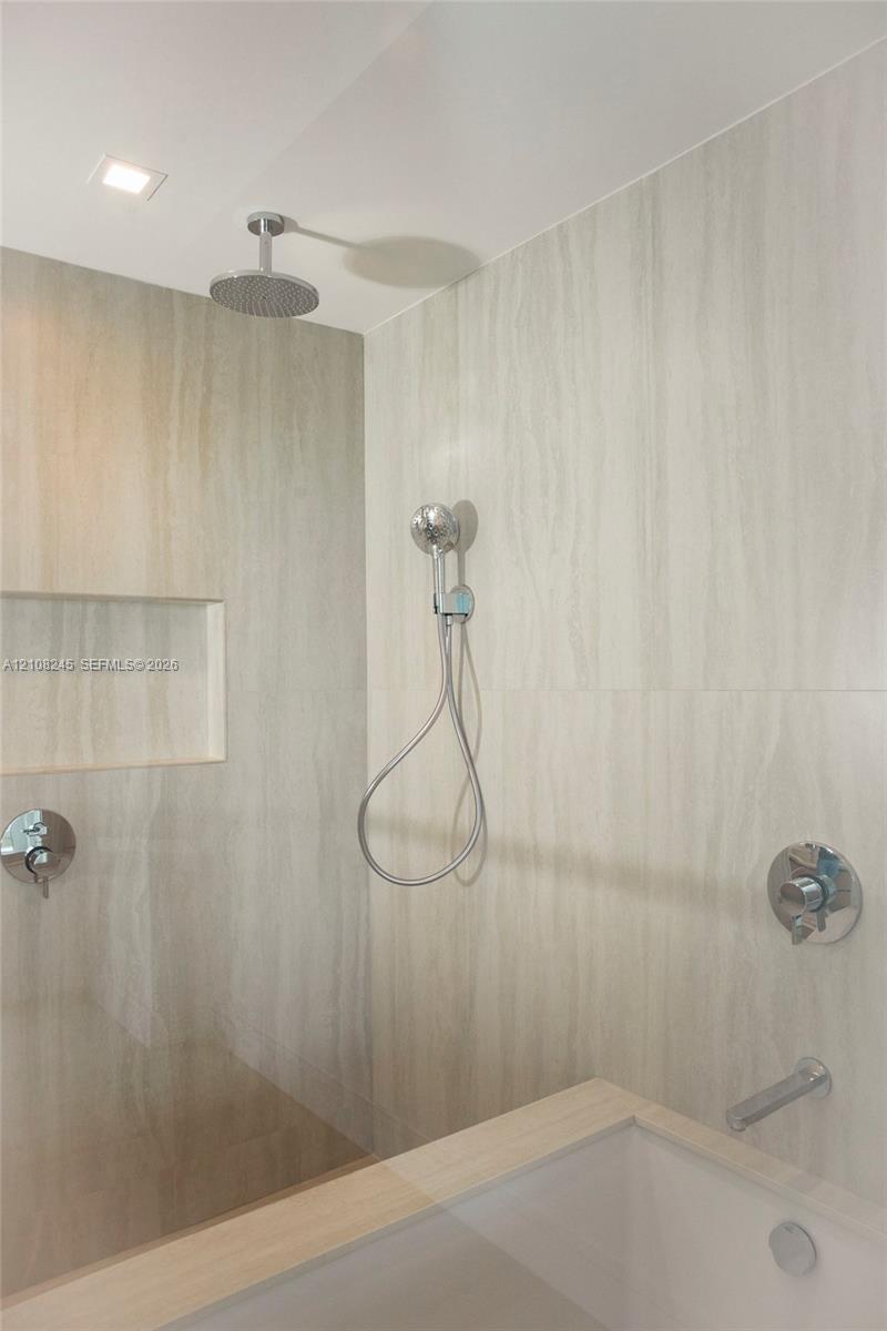 Walk-in shower featuring large-format tile surround, recessed wall niche, chrome rainfall shower head, handheld sprayer, and polished chrome hardware