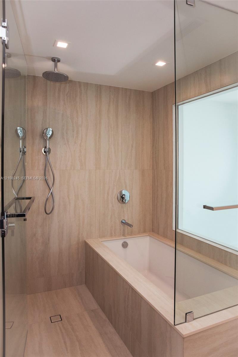 Walk-in shower and bathtub combination featuring floor-to-ceiling stone-tiled walls, glass enclosure, polished chrome rainfall shower head with handheld attachment, and recessed lighting