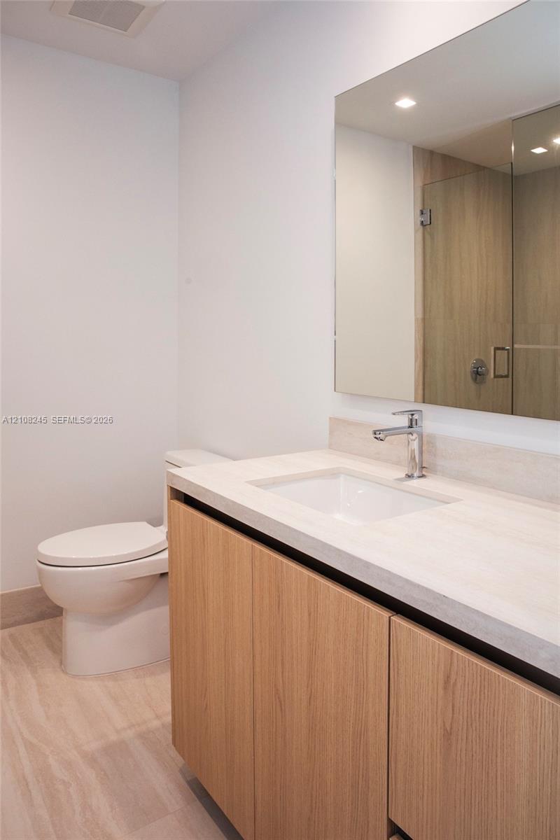 Bathroom featuring a wood-finish vanity with a light-toned countertop, integrated rectangular sink, polished chrome faucet, and a frameless wall mirror