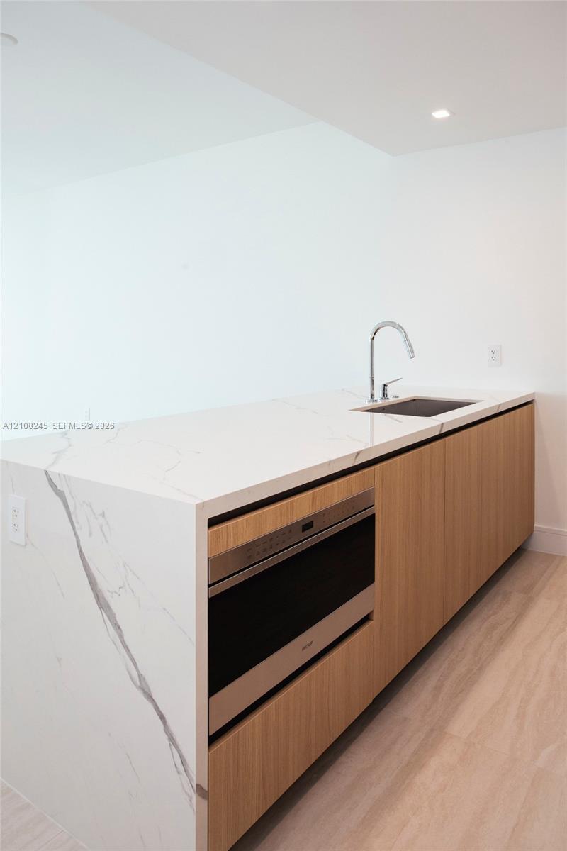 Kitchen island featuring a waterfall countertop with integrated sink and chrome pull-down faucet, flat-panel wood cabinetry, and a built-in microwave oven