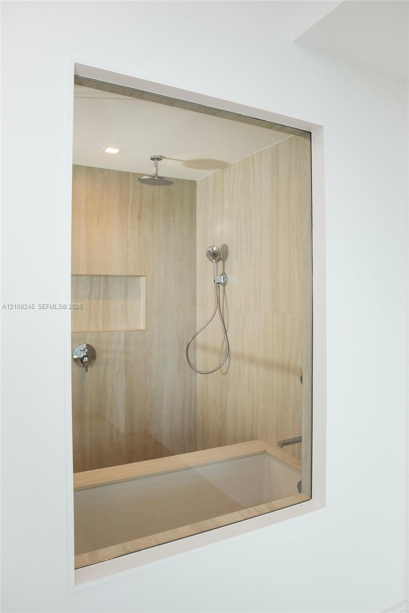 Walk-in shower enclosure featuring floor-to-ceiling neutral stone-finish tile, rainfall showerhead, handheld shower attachment, integrated wall niche, and glass panel partition