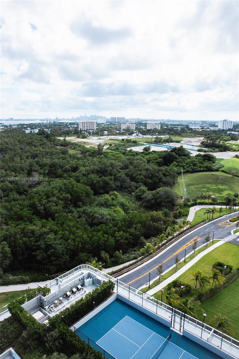 Aerial perspective featuring a hard-surfaced tennis court, rooftop patio area, metal perimeter railing, and surrounding greenery