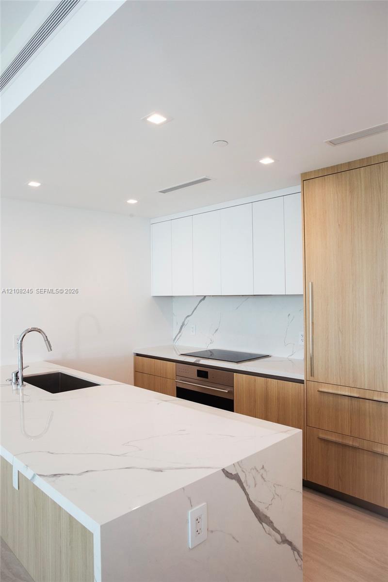 Kitchen featuring a central island with a stone countertop and waterfall edge, custom wood-finish cabinetry, white upper cabinets, integrated stainless steel appliances, and a stone slab backsplash