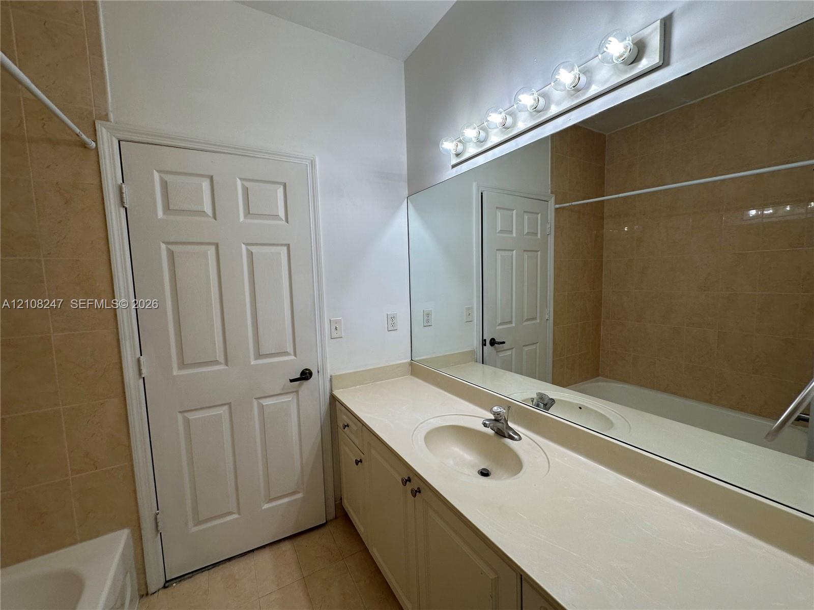 Full-width wall mirror, vanity cabinet with integrated countertop and sink, six-bulb light fixture, and floor-to-ceiling tiled shower enclosure
