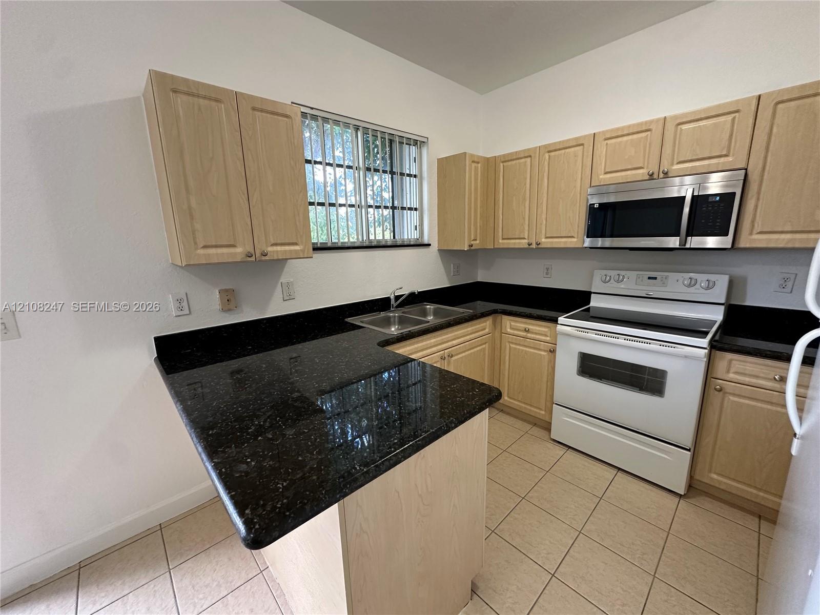 Kitchen featuring light-finish cabinetry, dark solid-surface countertops, dual-basin stainless steel sink, over-the-range microwave, and ceramic tile flooring
