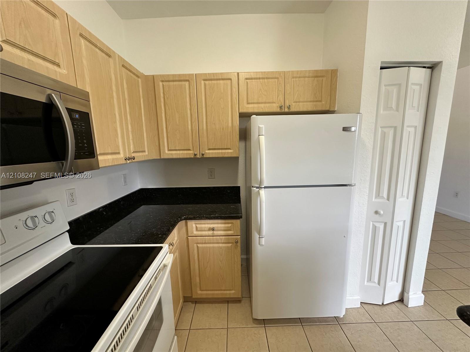 Kitchen featuring light-finish cabinetry, dark stone-style countertops, white appliance suite, and ceramic tile flooring
