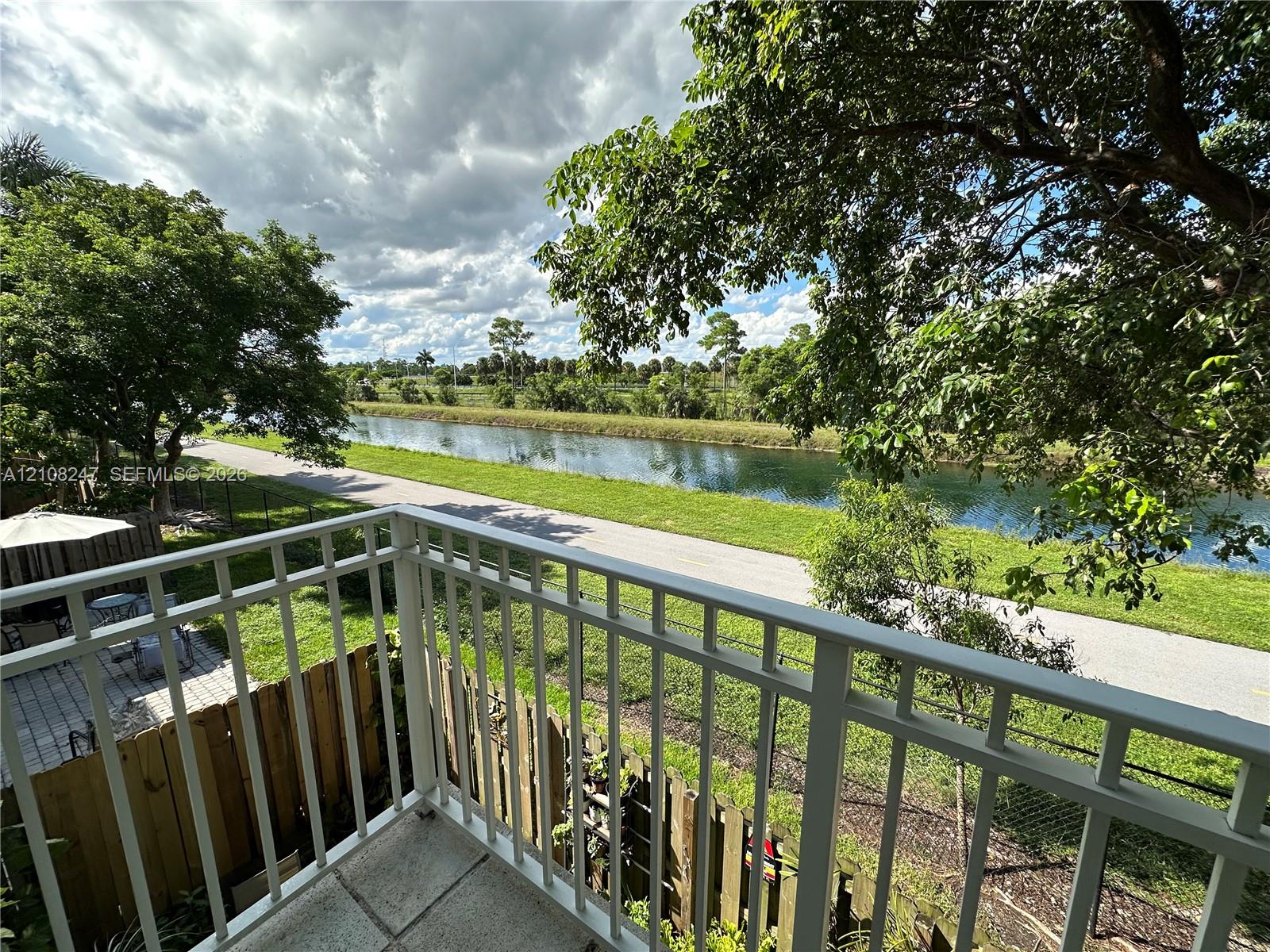 Elevated balcony with a white metal railing, paved patio area, wood fencing, and water frontage