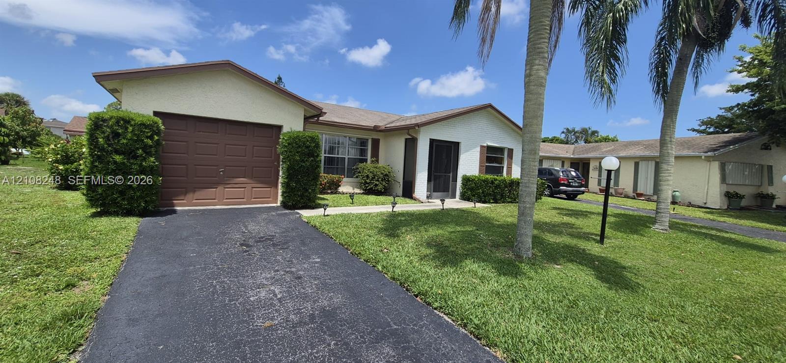 Single-story residence featuring a light-colored stucco exterior, attached garage with a solid-panel door, paved driveway, screen-enclosed entryway, and mature palm trees