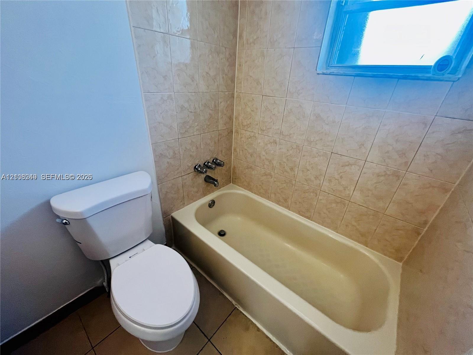 Bathroom featuring a porcelain tub, neutral ceramic wall tile, a window, and a standard-height toilet