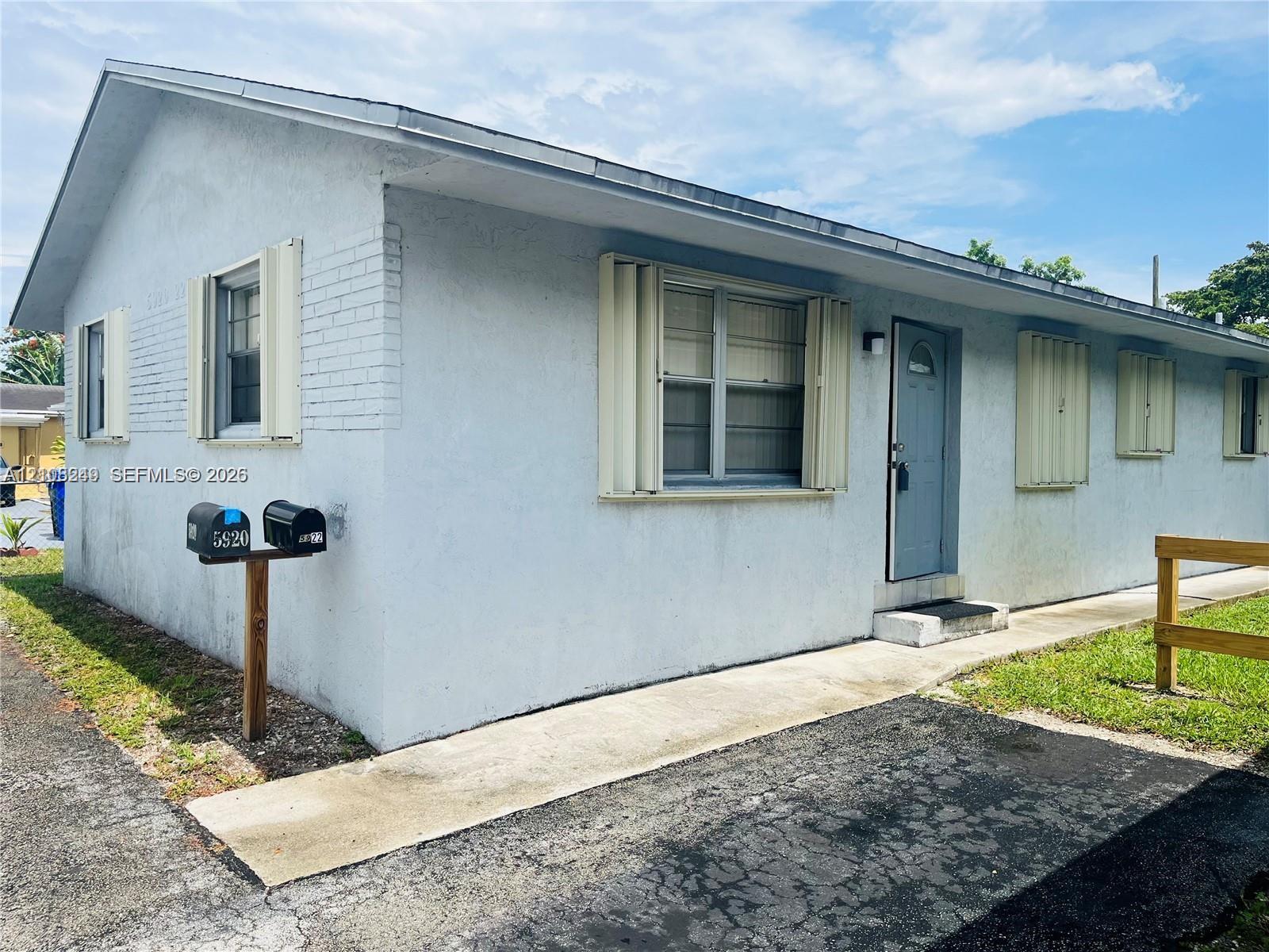 Stucco exterior featuring accordion hurricane shutters, a concrete walkway, and paved surface area