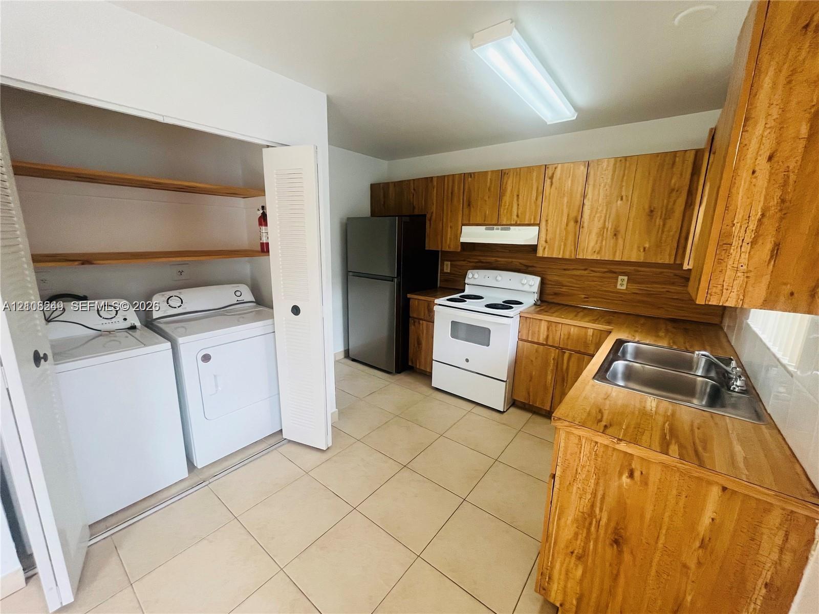 Kitchen featuring wood-finish cabinetry, neutral tile flooring, stainless steel refrigerator, white electric range, and a double-basin stainless steel sink
