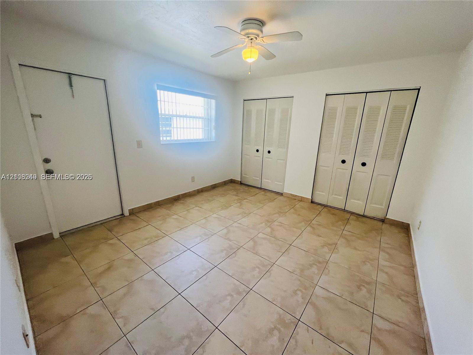 Room featuring large-format ceramic tile flooring, two sets of louvered bi-fold closet doors, a single-hung window, and a central ceiling fan with integrated lighting