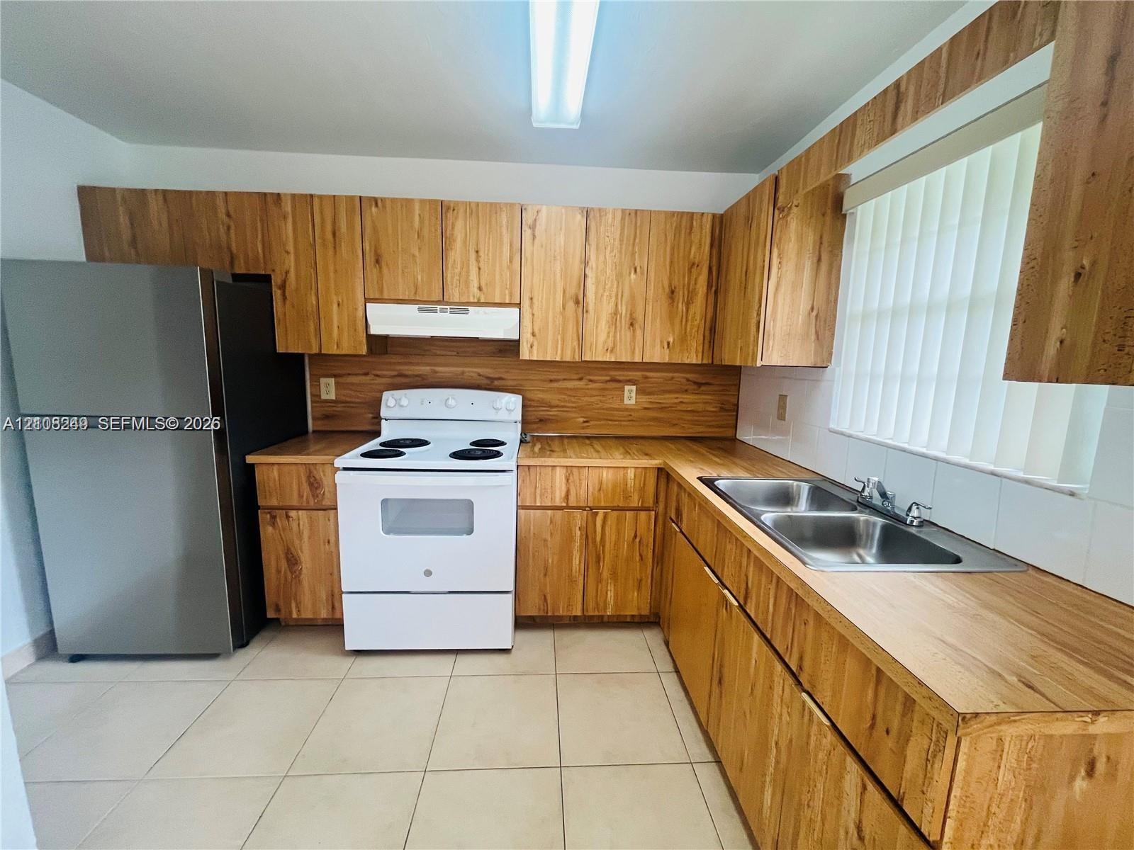 Kitchen featuring wood-finish cabinetry, wood-finish countertops, tile flooring, a dual-basin stainless steel sink, and stainless steel refrigerator