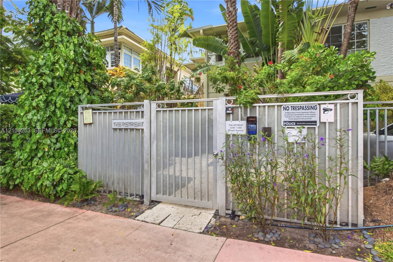 Gated entrance featuring metallic fencing, a secure access keypad, and established tropical landscaping