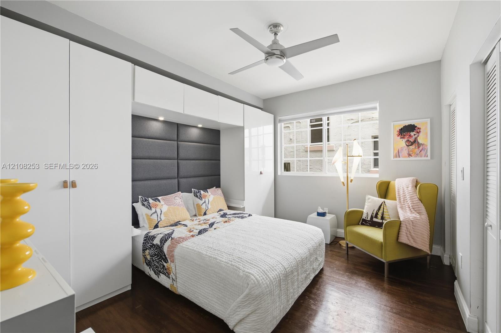 Room featuring dark wood-finish flooring, built-in white cabinetry, integrated recessed lighting, a charcoal grey upholstered headboard, and a multi-blade ceiling fan